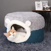 Panier Chat Coussin & Grotte – le Refuge Douillet pour Votre Félin ! Chat sortant sa tête de la grotte, sur tapis moelleux gris avec une table et un livre DESIGN HOTELS à l'arrière droite