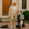 Poussette pour chat à deux étages avec sacs de rangement, pratique et confortable pour les promenades en extérieur. Une femme 60 ans cheveux blanc a une poussette pliée, ses deux chats noir et blanc sont présents devant leur maison.
