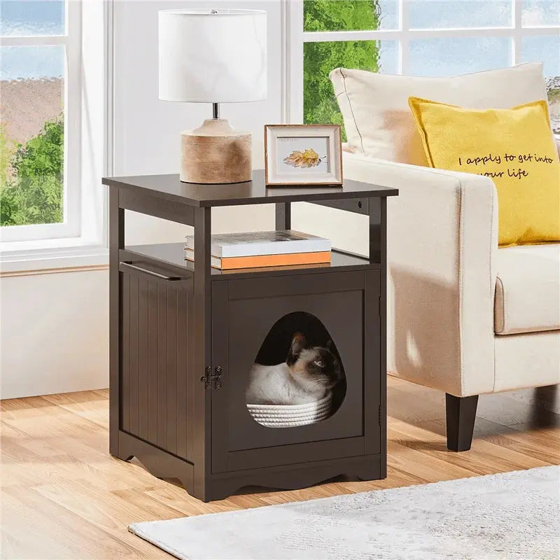 Meuble Litière Chat en Forme de Table d'Appoint avec un chat gris et noir à l'intérieur une lampe un cadre et deux livre au dessus un canapé à droite et des fenêtres derrière 