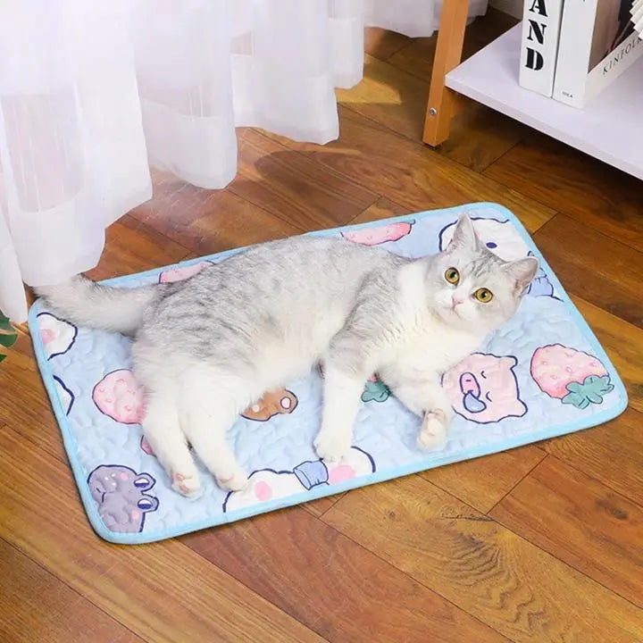 Tapis Rafraichissant Chat avec Motifs d'Animaux et de Fraises avec un chat gris et blanc allongé dessus avec des rideaux blanc derrière et un meuble à droite avec des livres posés dessus
